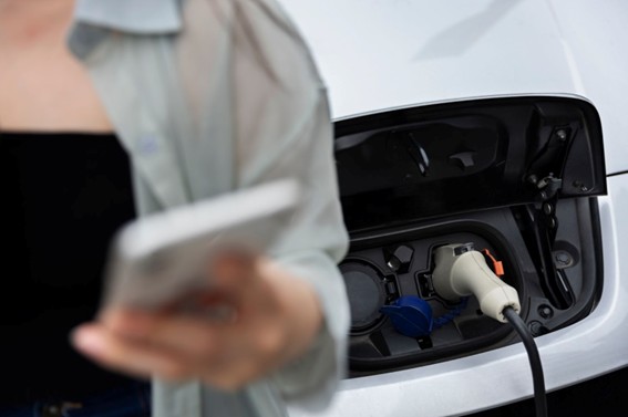 Persona usando el celular frente a un auto eléctrico que está siendo cargado mediante un enchufe conectado a su puerto de carga.