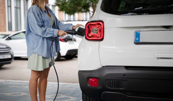 Mujer joven conectando un vehículo eléctrico blanco en una estación de carga urbana, simbolizando la movilidad sostenible.
