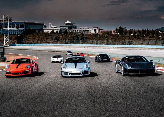 Varios autos superdeportivos de lujo, incluyendo Porsche y Ferrari, avanzan en una pista de carreras con un edificio de fondo y cielo nublado.