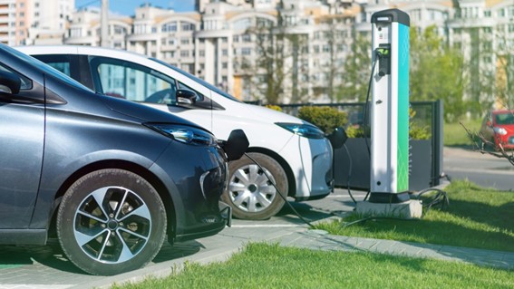 Dos autos eléctricos conectados a una estación de carga en una ciudad moderna, sobre una vereda con césped.