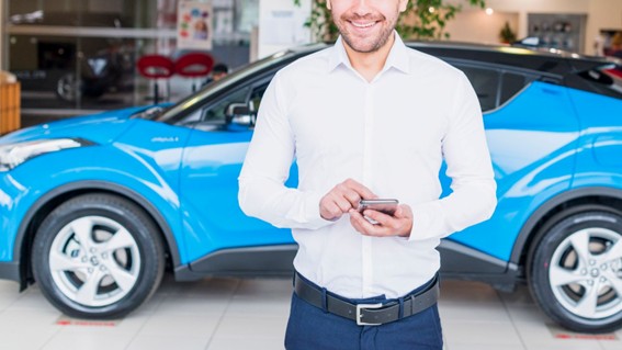 Hombre sonriente con camisa blanca usando el celular frente a un auto azul en una concesionaria.