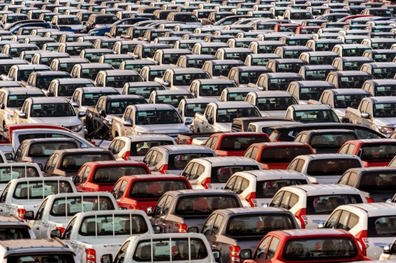 Gran cantidad de autos nuevos, blancos, rojos y grises, estacionados en filas simétricas en un predio industrial, representando el almacenamiento masivo previo a su distribución comercial.