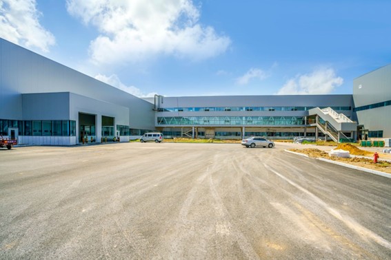 Edificio industrial moderno con amplias áreas de acceso y una pasarela elevada, rodeado de un espacio en construcción y cielo parcialmente nublado.