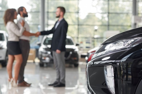 Pareja recibe su nuevo automóvil de un asesor de ventas en una concesionaria, con autos en exhibición y un vehículo negro en primer plano.