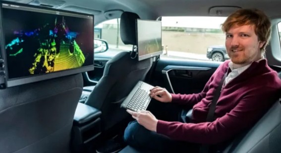 Hombre sentado en el asiento trasero de un auto, usando un teclado inalámbrico frente a dos pantallas con visualización 3D colorida.