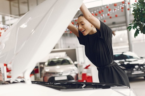 Mujer sonriente levantando el capó de un coche blanco en un concesionario con autos en exhibición al fondo.
