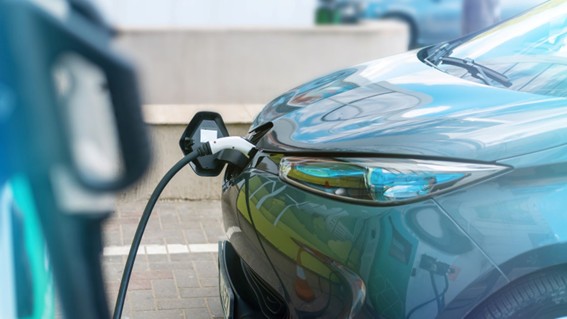 Vehículo eléctrico recibiendo carga en una estación pública, con el cable conectado en su parte frontal y fondo urbano desenfocado.