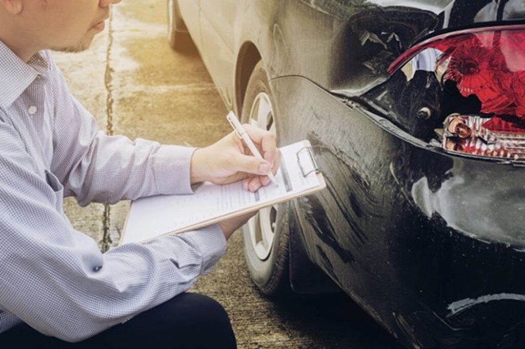 Hombre tomando notas en una planilla mientras inspecciona los daños en la parte trasera de un automóvil negro accidentado.