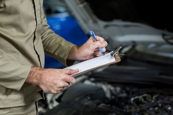 Persona escribiendo en un portapapeles frente a un auto con el motor descubierto durante una revisión técnica.