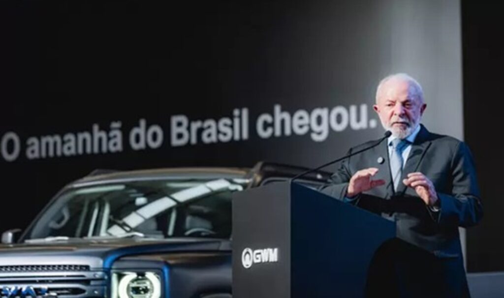 Presidente de Brasil en un evento de GWM, hablando frente a un podio con un automóvil eléctrico exhibido y el lema "O amanhã do Brasil chegou" proyectado en el fondo.